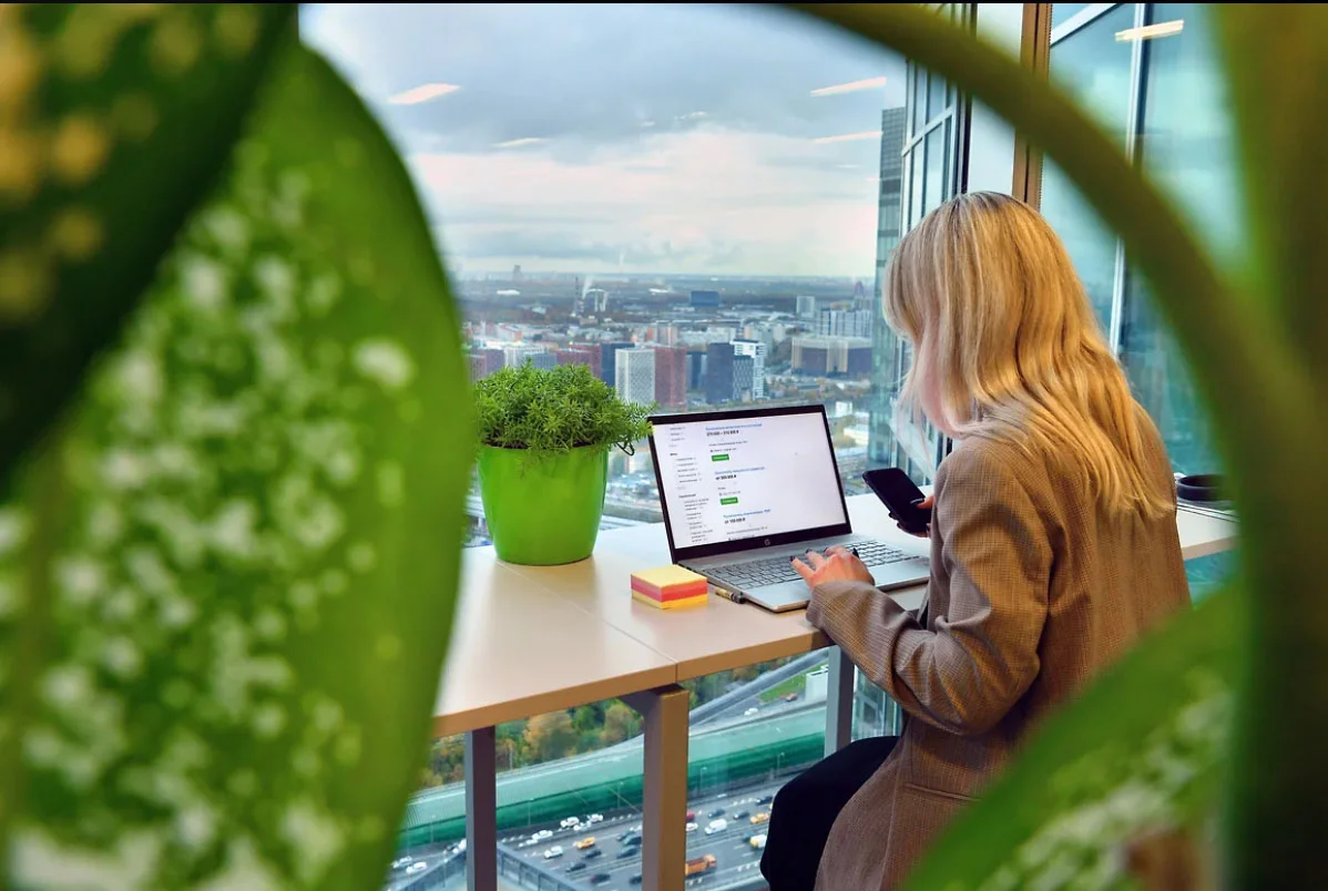 девушка с ноутбуком и смартфоном изучает рынок труда сидя у большого окна высотного здания