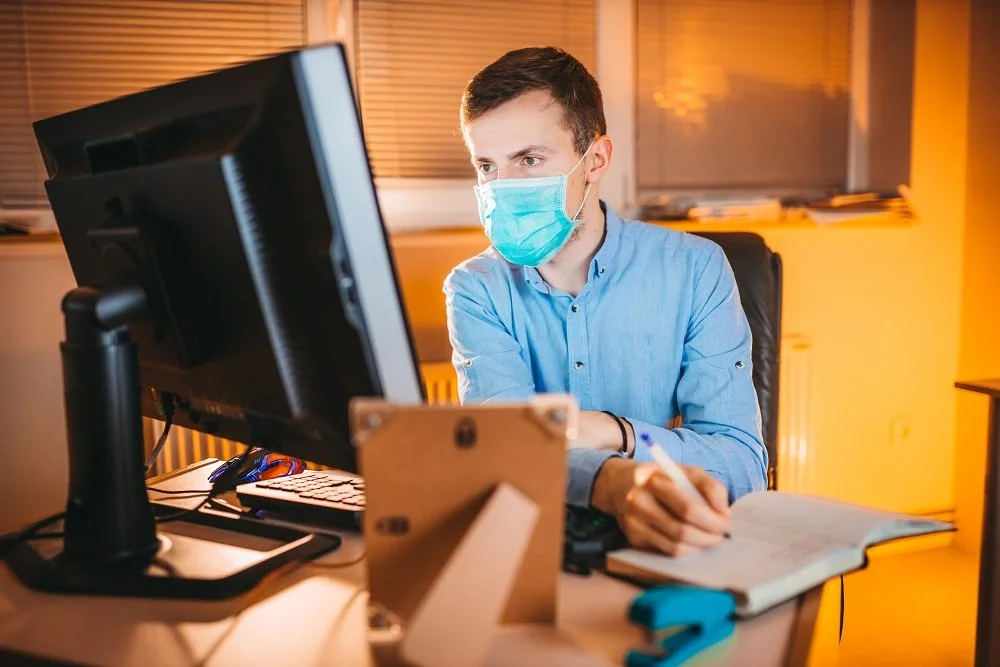 офисный сотрудник на рабочем месте за ПК с ручкой и записной книжкой в защитной маске