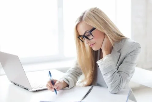 Девушка бухгалтер на первичную документацию сидит за столом с ноутбуком и пишет в журнале