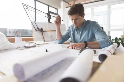 Мужчина инженер-проектировщик ВК НВК сидит за столом в офисе с чертежами и держит в руках карандаш и треугольник