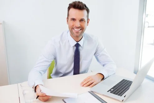 Мужчина менеджер по продажам электрооборудования сидит за столом с ноутбуком и держит в руке документ