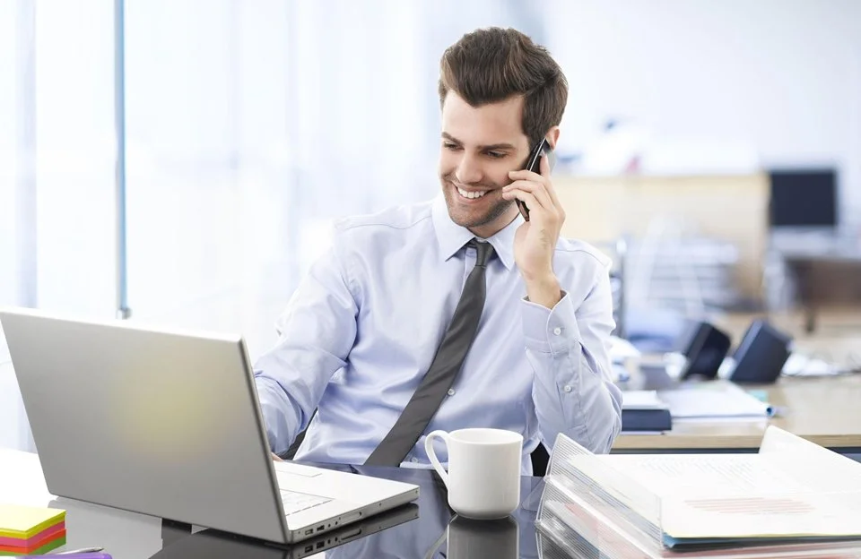 Мужчина менеджер по продажам сидит в офисе за столом с ноутбуком и разговаривает по телефону