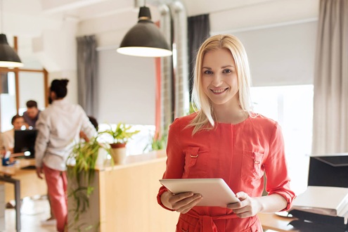 Девушка менеджер по продажам мебели в красном платье стоит в офисе на фоне сотрудников и держит планшет