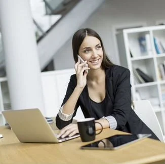 Девушка менеджер по продажам сидит за столом с ноутбуком, чашкой и планшетом и разговаривает по телефону