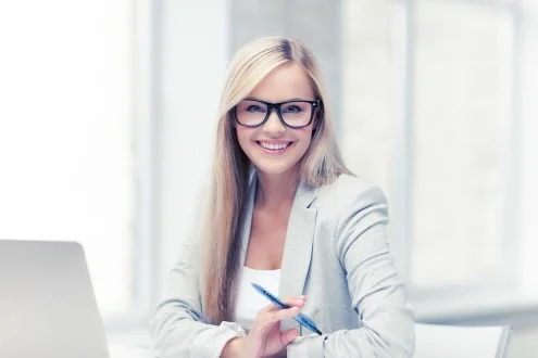 Девушка младший юрист в очках сидит в офисе за столом с ноутбуком и ручкой