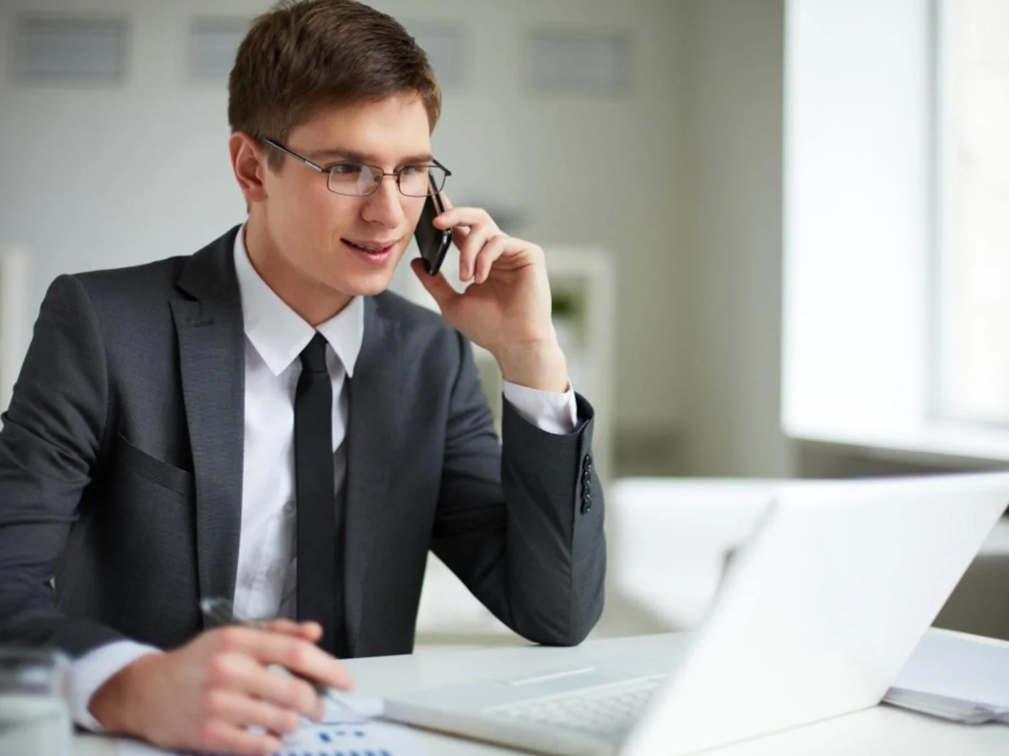 Мужчина менеджер по продажам в очках сидит за столом с ноутбуком и разговаривает по телефону