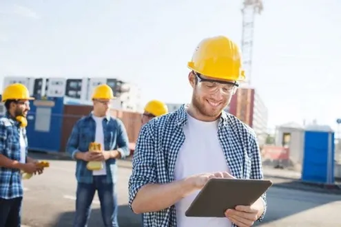 Мужчина начальник участка в желтой каске и очках работает с планшетом на фоне рабочих