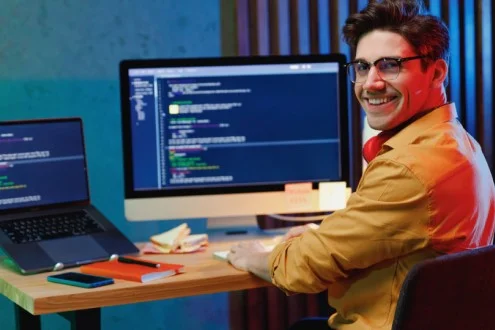 Молодой разработчик 1С работает за столом с ноутбуком и монитором