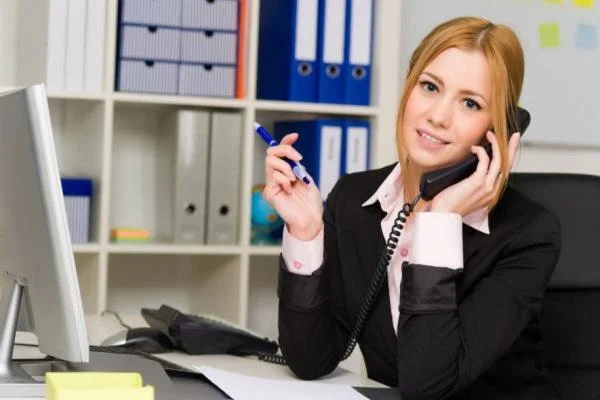Девушка референт общается по телефону за столом в офисе и держит в руке ручку