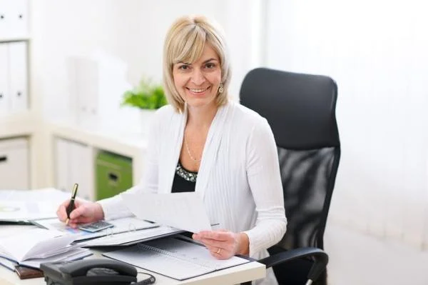 Женщина руководитель отдела продаж сидит за столом в кресле и делает запись в журнал