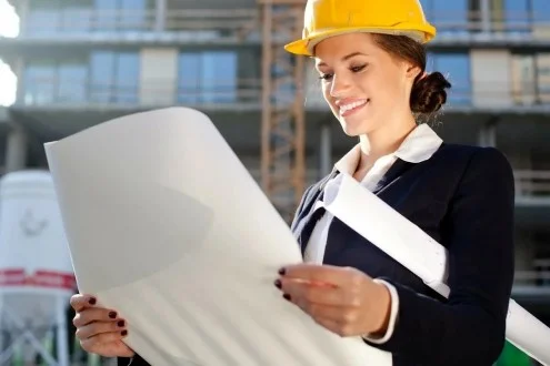 Девушка сметчик в каске стоит на фоне стройки и смотрит на чертеж