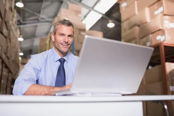 Мужчина Специалист WMS сидит за столом с ноутбуком на фоне стеллажей