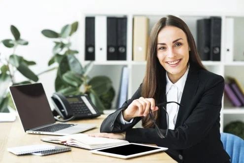 Девушка заместитель главного бухгалтера сидит за столом с ноутбуком, планшетом и записной книжкой